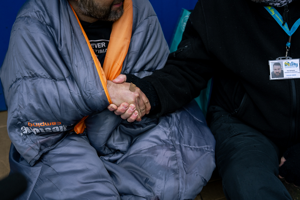 A homeless man in a sleeping bag, reaching out to shake hands.